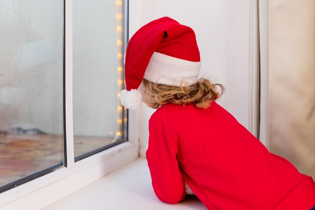 Natal Criança Duende no Chapéu de Papai Noel, duende. Feliz Natal e boas festas. menina sentada perto da janela e olhando para a floresta de inverno.