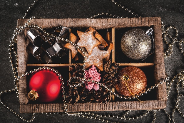 Natal, decoração do feriado. Bolas de árvore de Natal, guirlanda, estrelas de gengibre, molde de biscoitos, arcos para presentes, cones de abeto em caixa de madeira na mesa de pedra preta, vista superior