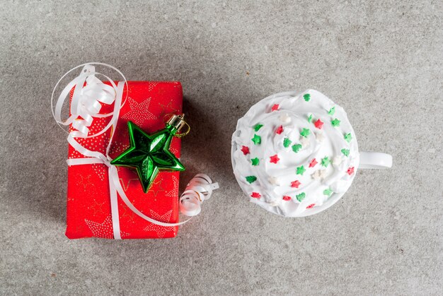 Natal e. uma caixa de presente de natal em papel vermelho e copo para café ou chocolate quente, com chantilly e decoração de estrelas doces, na mesa de pedra cinza, vista superior