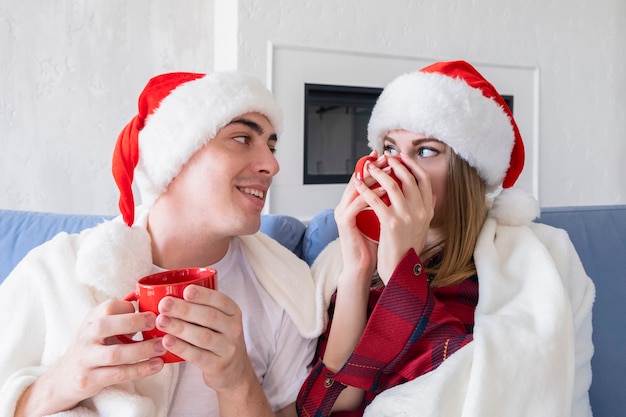 Natal em casa conceito. Foto de perfil de vista lateral de duas pessoas adoráveis e despreocupadas sentadas em um sofá aconchegante ou sofá na sala de estar segurando xícaras vermelhas olhando uma para a outra com um sorriso