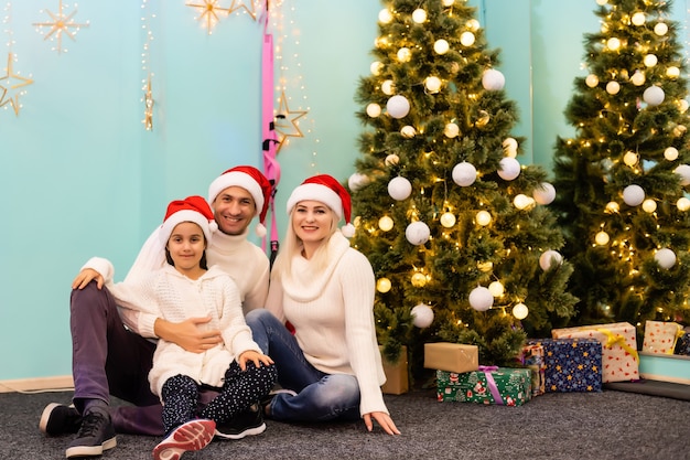 Natal. Família. Felicidade. pai, mãe e filha com gorros de Papai Noel olhando para a câmera e sorrindo no chão em casa