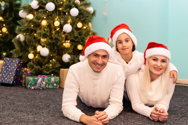 Natal. Família. Felicidade. pai, mãe e filha com gorros de Papai Noel olhando para a câmera e sorrindo no chão em casa