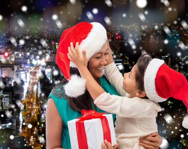 natal, feriados, celebração, família e conceito de pessoas - feliz mãe e filha em chapéus de ajudante de papai noel com caixa de presente sobre fundo