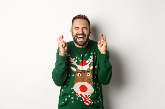 Natal, feriados e comemorações. Cara esperançoso e animado fazendo um desejo, mantenha os dedos cruzados para boa sorte e sorrindo, em pé sobre um fundo branco.