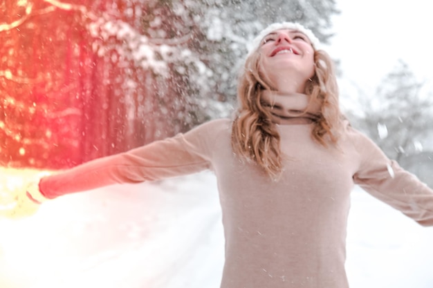 Natal, feriados e conceito de temporada. jovem mulher feliz soprando neve na natureza da floresta de inverno. luvas e gorro de malha de roupas quentes. fundo de paisagem de floresta de inverno