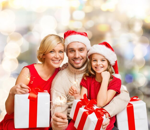 natal, feriados, família e conceito de pessoas - feliz mãe, pai e filha em chapéus de ajudante de papai noel com caixas de presente sobre fundo de luzes