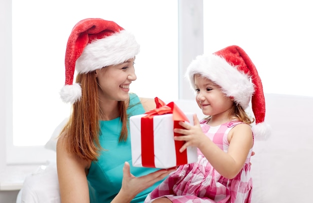 natal, feriados, família, infância e conceito de pessoas - feliz mãe e menina em chapéus de papai noel com caixa de presente em casa