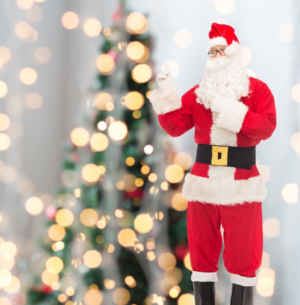 natal, feriados, gesto e conceito de pessoas - homem fantasiado de papai noel apontando dedos sobre fundo de luzes de árvore