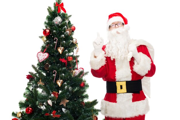 natal, feriados, gesto e conceito de pessoas - homem fantasiado de papai noel com saco e árvore de natal apontando o dedo para cima