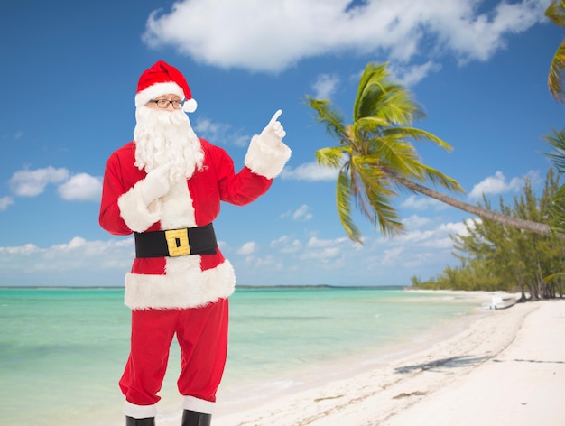 natal, feriados, gesto, viagens e conceito de pessoas - homem fantasiado de papai noel apontando dedos sobre fundo de praia tropical