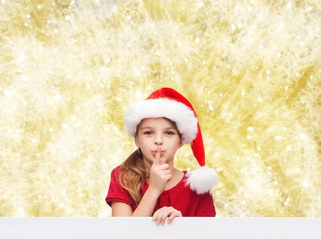 natal, feriados, infância e conceito de pessoas - menina sorridente no chapéu de ajudante de papai noel com o dedo nos lábios sobre fundo de luzes amarelas