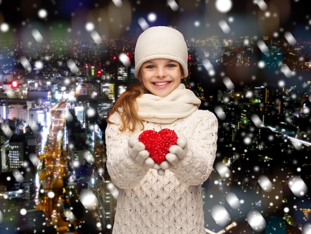 natal, feriados, infância, presentes e conceito de pessoas - sonhando menina em roupas de inverno com coração vermelho sobre o fundo da cidade de noite de neve