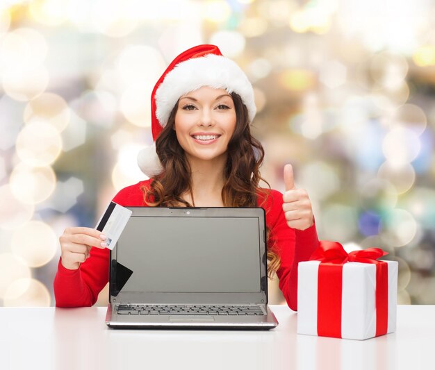 natal, feriados, tecnologia e conceito de compras - mulher sorridente no chapéu de ajudante de papai noel com cartão de crédito, caixa de presente e computador portátil mostrando os polegares para cima gesto sobre fundo de luzes