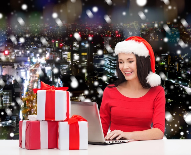 natal, feriados, tecnologia e conceito de pessoas - mulher sorridente no chapéu de ajudante de papai noel com caixas de presente e computador portátil sobre o fundo da cidade de noite de neve