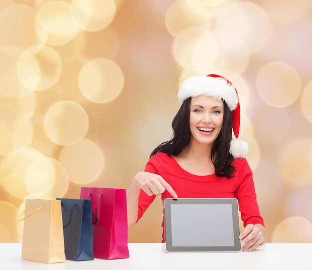 natal, feriados, tecnologia, publicidade e conceito de pessoas - mulher sorridente no chapéu de ajudante de papai noel com sacolas de compras e computador tablet pc sobre fundo de luzes bege