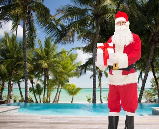 natal, feriados, viagens e conceito de pessoas - homem fantasiado de papai noel com caixa de presente sobre praia tropical e fundo de piscina