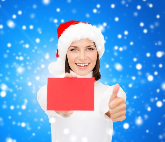 Foto natal, holdays, pessoas, propaganda e conceito de venda - mulher feliz no chapéu de ajudante de papai noel com cartão vermelho em branco, mostrando os polegares para cima gesto sobre fundo azul nevado