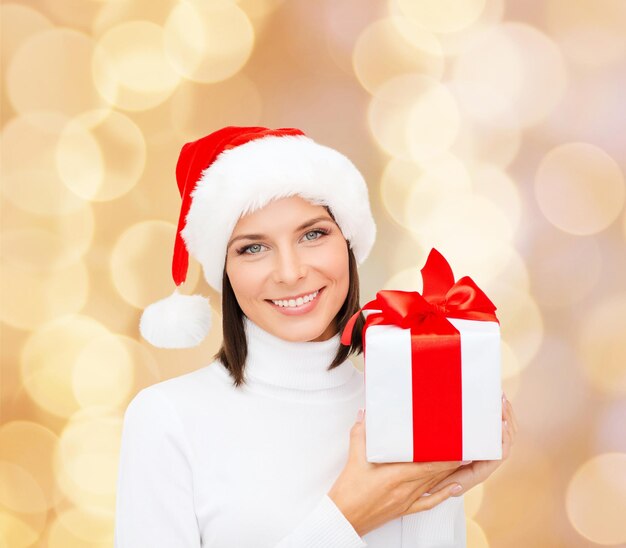 natal, inverno, felicidade, feriados e conceito de pessoas - mulher sorridente no chapéu de ajudante de papai noel com caixa de presente sobre fundo de luzes bege