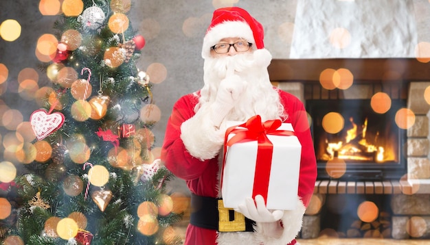 natal, inverno, feriados e conceito de pessoas - homem fantasiado de papai noel com caixa de presente e árvore fazendo gesto de silêncio sobre lareira em casa e fundo de luzes