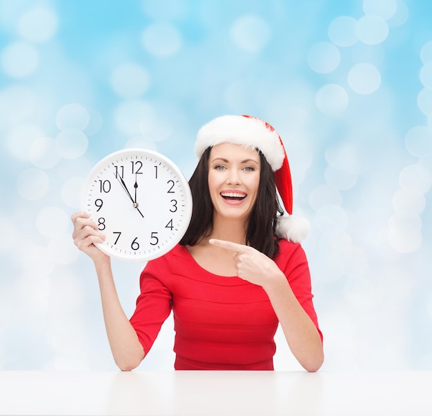 natal, inverno, feriados, tempo e conceito de pessoas - mulher sorridente com chapéu de ajudante de papai noel e vestido vermelho com relógio sobre fundo de luzes azuis