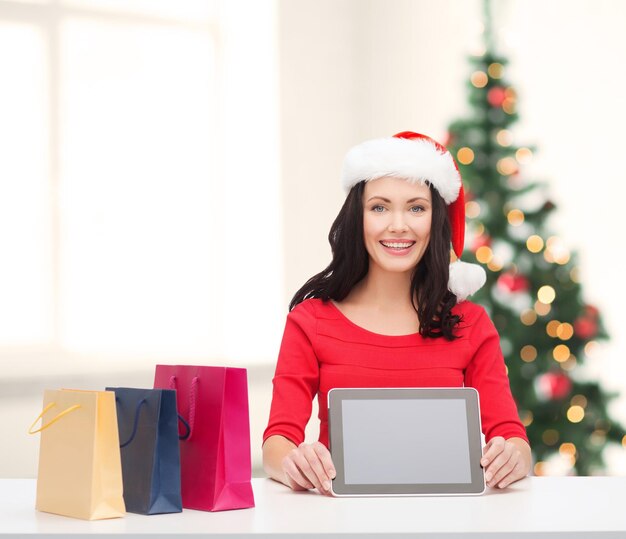 natal, natal, eletrônicos, conceito de gadget - mulher sorridente com chapéu de ajudante de Papai Noel com tela em branco do tablet pc e sacolas de compras