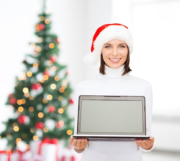 natal, tecnologia, férias de inverno e conceito de pessoas - mulher sorridente no chapéu de ajudante de papai noel com computador portátil de tela em branco sobre a sala de estar e o fundo da árvore de natal