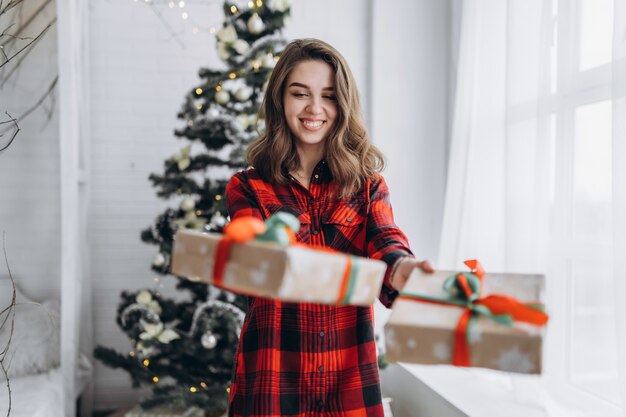 Natal. Uma linda mulher com camisa e meias andando em casa com caixa de presente de natal, árvore de natal atrás