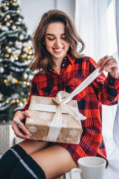 Natal. Uma linda mulher de camiseta e meias sentada no parapeito da janela com uma caixa de presente de Natal, a árvore de Natal atrás