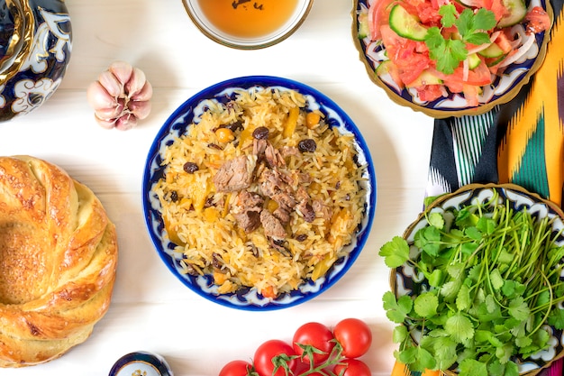 Nationaler usbekischer Pilaw mit Fleisch, Achichuk-Salat von Tomaten, Gurken, Zwiebeln in Teller mit traditionellem Muster, Koriander, Kirschtomaten, Knoblauchbrot-Tortilla - Patir auf weißem Holztisch Draufsicht