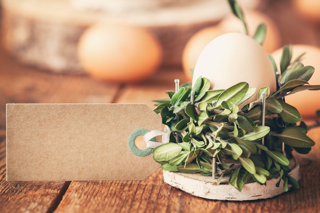 Natürliche Eier im Korb von Bündelpflanze und leerem Etikett für Text auf Holztisch im sonnigen Morgen. Umweltfreundliches Osterkonzept.