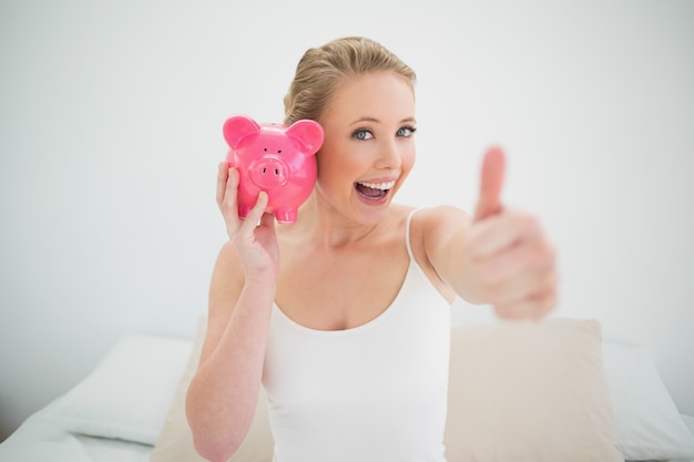 Natürliche glückliche Blondine, die Sparschwein beim Sitzen auf Bett hält