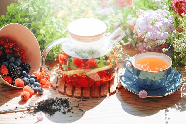 Natürlicher aromatischer Früchtetee in einer transparenten Teekanne und in einer Tasse zwischen Beeren und Blumen wärmender aromatischer Tee mit einem tiefen Aroma von Beeren und Wildblumen