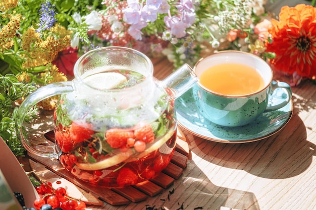 Natürlicher aromatischer Früchtetee in einer transparenten Teekanne und in einer Tasse zwischen Beeren und Blumen wärmender aromatischer Tee mit einem tiefen Aroma von Beeren und Wildblumen