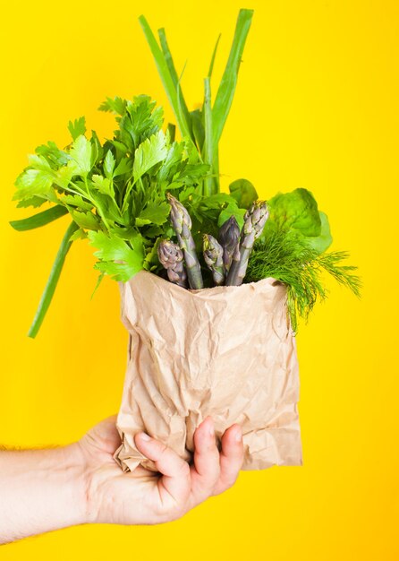 Natürliches gesundes Lebensmittel auf gelbem Hintergrund. Spargel und Grün für Salat in einer Papiertüte in der Hand, Ökoladen