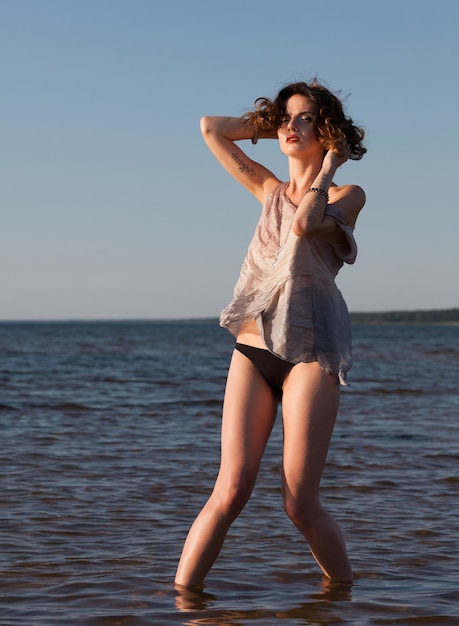 Natur, Schönheit, Jugend und gesundes Lebensstilkonzept. Attraktive junge halbnackte Frau in einem Neoprenanzug posiert vor dem Meereshintergrund. Abendlicht mit tiefen Schatten und Farbverschiebung