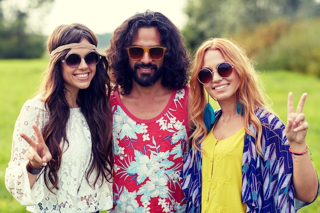 Natur-, Sommer-, Jugendkultur-, Gesten- und Menschenkonzept - lächelnde junge Hippie-Freunde mit Sonnenbrillen, die ein Friedenshandzeichen auf der grünen Wiese zeigen