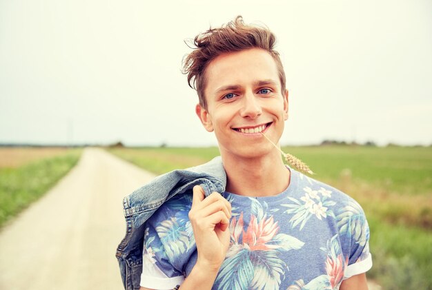 Foto natur-, sommer-, jugendkultur- und menschenkonzept - lächelnder junger hippie-mann auf der landstraße, der roggenspitze kaut