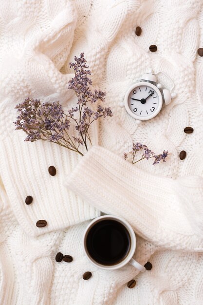 Foto naturaleza muerta inspiradora: reloj despertador, taza de café y flores secas en un acogedor suéter blanco. tiempo para descansar y relajarse. vista superior y vertical