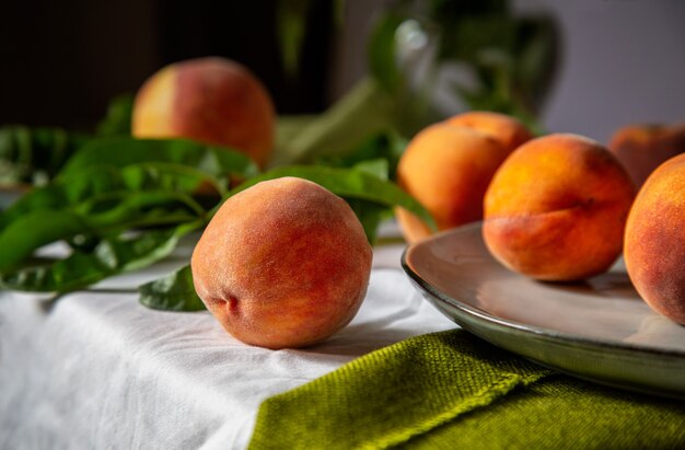 Naturaleza muerta Melocotones fruta. Melocotones en la mesa de la cocina junto a la ventana. Frutas de melocotón de comida de minimalismo rústico con hojas sobre tabla de madera sobre mantel. Cosecha de melocotones jugosos maduros.