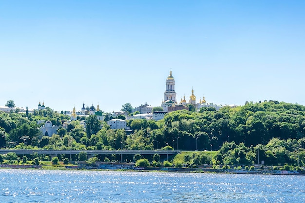 Natureza cênica viagem paisagem urbana rio Dnipro com água azul e histórico mosteiro do templo da igreja Lavra no dia ensolarado de verão em KievUcrâniaVista de dentro do rio da margem verde do parque da cidade com árvores