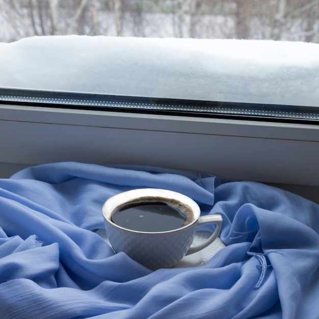 Natureza-morta aconchegante de inverno: uma xícara de café quente, um lenço azul no parapeito da janela contra o fundo de uma paisagem de neve