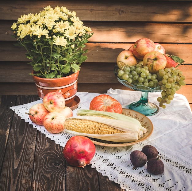 Natureza morta de outono em estilo rústico com maçãs, abóbora e milho, romã e figos em uma toalha de mesa branca com renda e flores amarelas de crisântemo