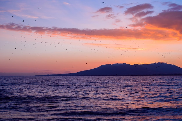 Natureza no período do crepúsculo no pôr do sol do mar sobre a montanha e um bando de pássaros voando Lindas nuvens pintadas na cor laranja Fundo da natureza