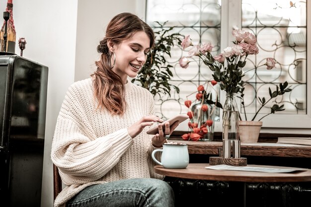 Navegación por Internet. Una mujer sonriente navegando por internet usando un teléfono inteligente mientras bebe cacao