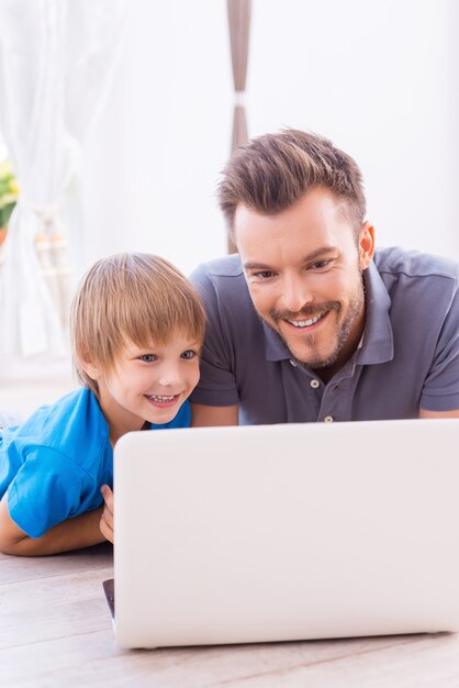 Navegando por la red juntos. Feliz padre e hijo mirando la computadora portátil y sonriendo mientras está acostado en el piso en casa