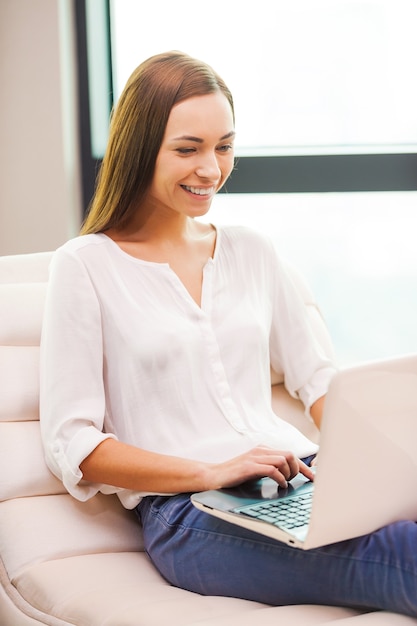 Navegar por la red en casa. Hermosa mujer joven que trabaja en la computadora portátil y sonriendo mientras está sentado en la silla en casa