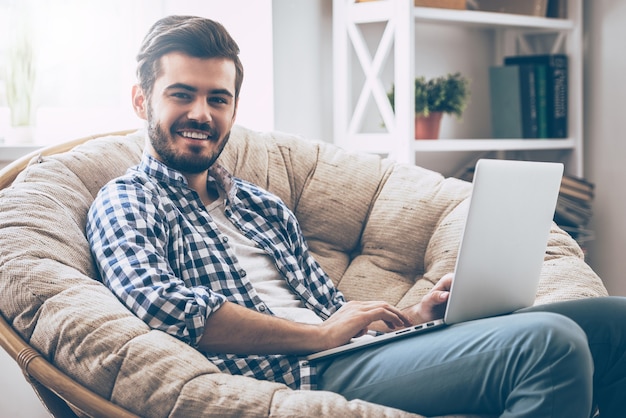 Navegar por la web en casa. Apuesto joven trabajando en una computadora portátil y sonriendo mientras está sentado en una silla grande y cómoda en casa