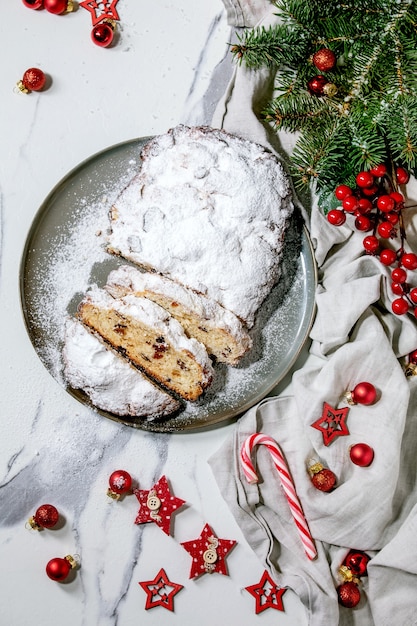 Navidad alemana casera tradicional hornear pan de tarta stollen en placa con adornos navideños y ramas de abeto sobre superficie de mármol blanco. Endecha plana