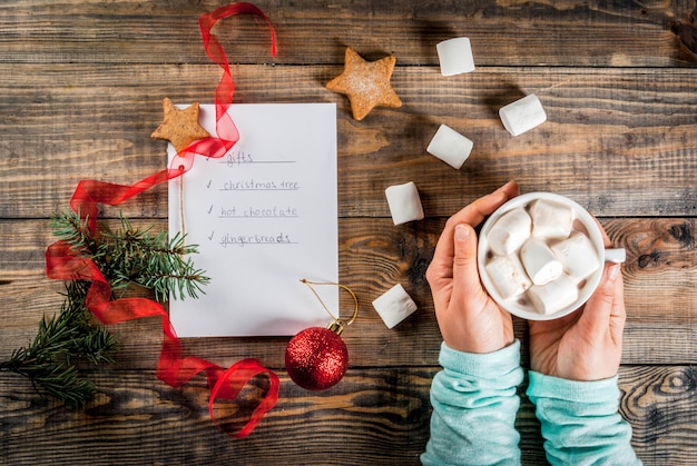 Navidad, año nuevo concepto. Mesa de madera, cuaderno con lista de tareas, las manos de la niña sostiene la taza de cacao, la bola de Navidad, el pino, la cinta roja, el malvavisco Vista superior copyspace