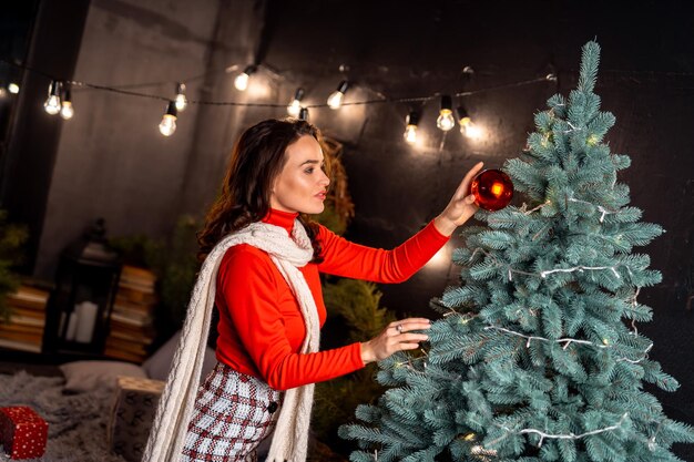 Navidad Año Nuevo concepto de vacaciones de invierno joven dama sonriente feliz decora el árbol de Año Nuevo posando en casa decorada festiva modelo vistiendo suéter rojo